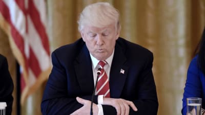 Donald Trump sitter vid ett bord med en mikrofon framför sig, med händerna knäppta och blicken vänd nedåt.