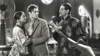 Kiinalaiseen perinneasuun pukeutunut mies (näyttelijä Boris Karloff) pitelee valtavaa miekkaa; häntä katsoo huolestuneen näköisenä eurooppalainen mies, jonka kainalossa on kiinalainen nainen. Kuva elokuvasta Fu Manchun naamio.