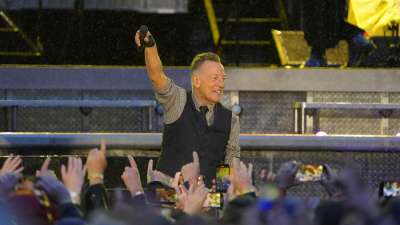 Bruce Springsteen står på scenen iklädd en väst över en skjorta och håller upp micken till en fullsatt arena.