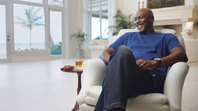 Michael Jordan sitter i en fotölj och skrattar, med ett whiskeyglas bredvid sig. 