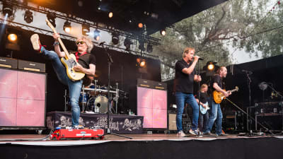 En musikgrupp spelar på en scen. En gitarrist har benet upp i luften trots att han spelar. Sångaren står i mitten och bredvid honom också två ytterligare gitarrister eller basister.