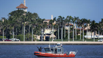 Kustbevakningens lilla röda båt med beväpnade vakter skyddar strandlilnjen till rpesidnetens lyxklubb som skymtar bakom palmerna.