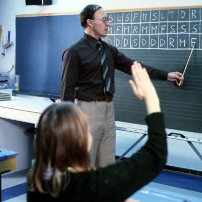 Opettaja liitutaulun edessä opettamassa solmisointi-käsimerkkejä (solmisointi,  käsimerkki). Oppilas tekee solmisointeja.