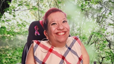 Anna Caldén småler stort mot kameran. Hon sitter i rullstol och bakgrunden är en grön trädgård.