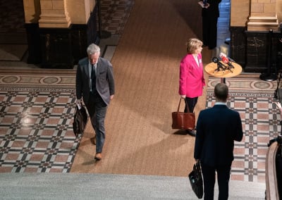En bild uppifrån riktad mot ett podium med en mikrofon. Anna-Maja Henriksson står intill mikrofonen, Anders Adlercreutz går iväg på väg ut ur bilden.
