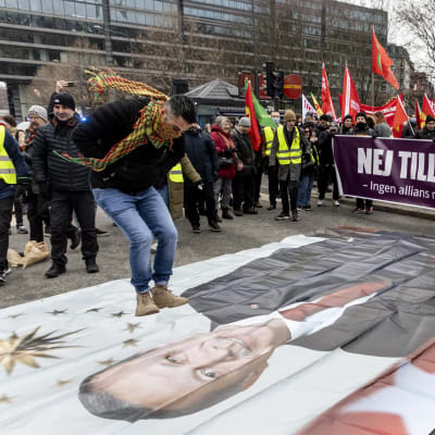 Ihmisiä mielenosoituksessa ulkona. Ihmisillä on Kurdistanin työväenpuolueen lippuja. Maassa on iso kangas, jossa Turkin presidentin Recep Tayyip Erdogan kuva. Mielenosoittaja hyppää kuvan Erdoganin kasvojen päällä.  
