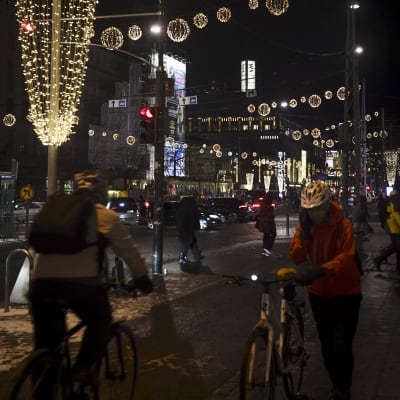 En person cyklar, en annan leder sin cykel. I bakgrunden syns glittrande julljus och en mörk stad.
