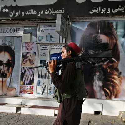 En talibansoldat bär på ett gevär. Han går förbi en affär där ansikten på kvinnor på raklamaffischer målats över.