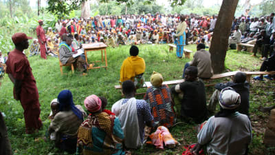 Iso ihmisjoukko on asettunut puoliympyrään ulos seuraamaan kyläkäräjia Ruandassa vuonna 2007. Keskellä pöytä, jonka ääressä istuu käräjien tuomareita ja pöydän edessä seisoo nainen. 