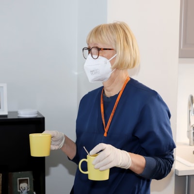 Lähihoitaja Anu Kuukka kävelee keltaiset kahvikupit käsissään keittiöstä olohuoneeseen. Käsissä suojakäsineet, kasvoilla maski., sininet työasu.