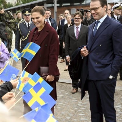 Kronprinsessan Victoria och prins Daniel på Sveaborg. Victoria i vinröd byxdress och Daniel i blå kostym ser båda glada ut och tittar på en grupp barn som viftar med handritade svenska flaggor.
