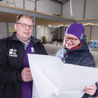 Kata Safety Oy:n yrittäjät Janne Rahunen ja Elina Rahunen. Heillä käsissään iso paperiarkki. Taustalla yrityshalli, tikapuut ja rakennustarvikkeita. 