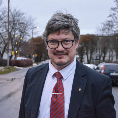 Medelålders man i kostym står på väg, tittar in i kameran med ett litet leende.