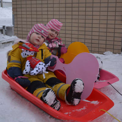 Lilja och Enni Latvakangas, två barn i vinterkläder och rosavitrandiga mössor, sitter i varsin pulka.