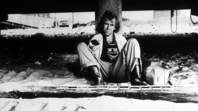 Mies istuu hiekalla nojaten betoniseen pylvääseen käsissään polaroid-kamera ja yksi valokuva, lisää kuvia on hiekalla hänen edessään.