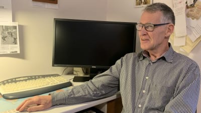 En äldre man med glasögon och rutig skjorta sitter vid ett skrivbord med en dator. 