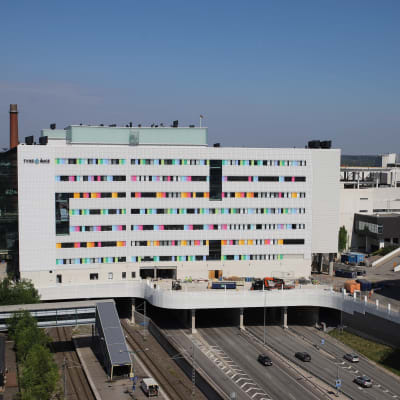 Valkoinen sairaalarakennus moottoritien päällä, värikkäitä yksityiskohtia ikkunoiden vieressä