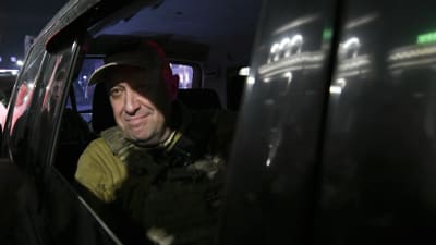 Jevgenij Prigozjin, klädd i skyddsväst, tittar ut genom fönstret i en mörk bil. Han ler och det är mörkt kring bilen.