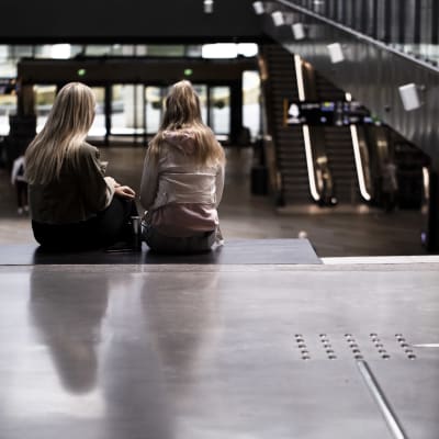 Två flickor med långt hår sitter med ryggen mot kameran på en bänk på köpcentret Tripla i Helsingfors.
