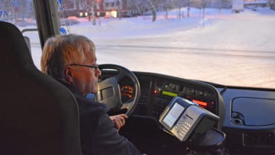 En man i medelåldern sitter bakom ratten på en buss. Det är mörkt i bussen, likaså utomhus. Ute synns en snötäckt gata.