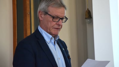 En medelålders man i glasögon tittar på ett papper. I bakgrunden syns en stor skärm.
