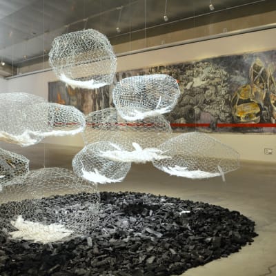 ett konstverk på en konstutställning med bollar gjorda av hönsnät som hänger från taket. I bollarna finns vita fjädrar. På golvet under bollarna finns en bädd av kol.