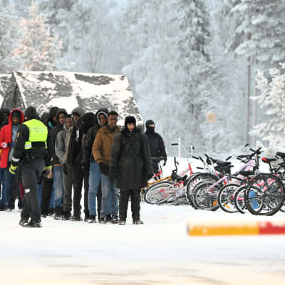 Joukko ihmisiä seisoo jonossa autopuomin edessä. Vieressä rivissä polkupyöriä lumisessa maisemassa. Rajavartija kävelee jonon vierellä.