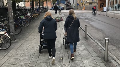Två kvinnor promenerar med varsin barnvagn i stadsmiljö.