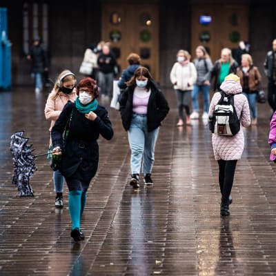 Många människor promenerar åt olika håll, med stora bruna dörrar i bakgrunden. Utanför Helsingfors järnvägsstation den 27 oktober 2020.