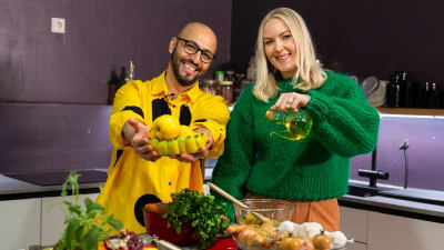 Mies keltaisessa paidassa ja nainen vihreässä paidassa katsoo kameraan. Edessä pöydällä eri ruoka-aineita, kuin sipulia, yrttejä ja oliiviöljyä.