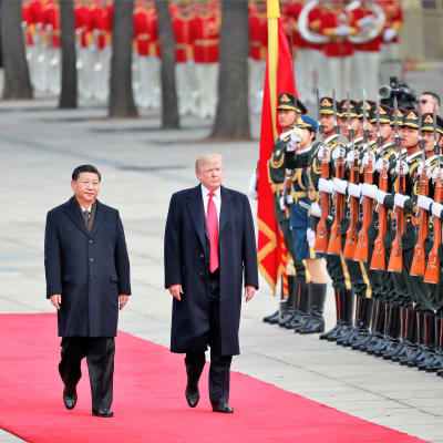 Presidentin Xi ja Trump kävelevät vakavina mustissa puvuissa ja päällystakeissa punaisella matolla kunniakaartin ohi. Xin takki on napitettu, Trumpin päällystakki on auki ja kirkkaanpunainen kravatti näkyy koko pituudeltaan.