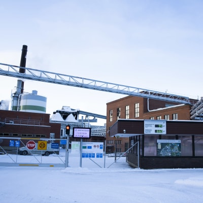 UPMn tehdas Valkeakoskella. Punatiilisiä rakennuksia, korkea piippu. Lunta maassa. Vartiokoppi ja aita edustalla. 