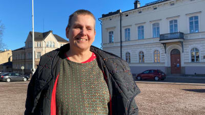 En korthårig kvinna står på ett tomt torg. Hon har jackan uppknäppt, solen i ansiktet och hon ler.