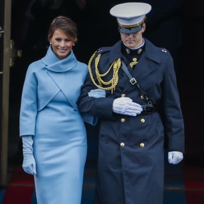 Melania Trump i ljusblå klänning och handskar i samma färg blir eskorterad av en soldat.