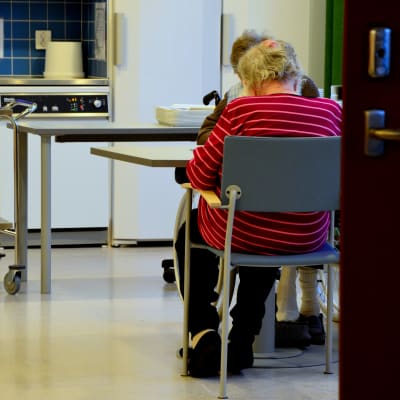 Ett par äldre personer sitter vid ett bord. I bakgrunden syns ett kök.