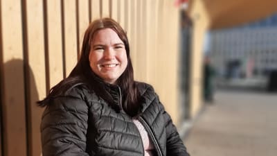 En kvinna med rakt långt mörkt hår och en svart dunjacka, där dragkeden är nerdragen, sitter i vårsolen på en stol vid en gul brädvägg.