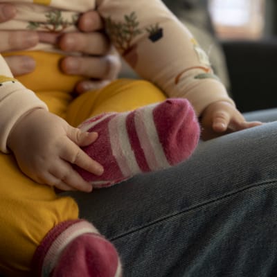 Vauva istuu aikuisen sylissä ja koskettaa omaa kantapäätään. Kuva rajautuu niin, ettei vauvan kasvoja näy.