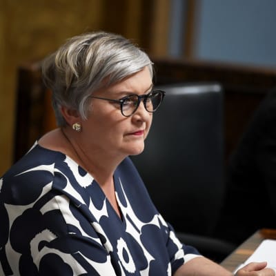 Anu Vehviläinen - En kvinna med kortklippt grått hår och svarta runda glasögon. Hon sitter i riksdagens plenisal iförd en blå marimekkoklänning.