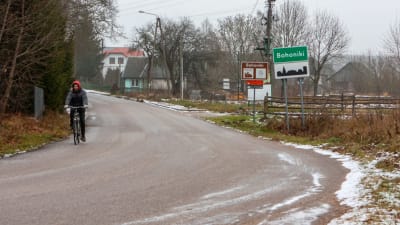 En asfaltväg leder in mot en samling äldre småhus. En man cyklar på vägen mot kameran. En skylt vid vägkanten med namnet "Bohoniki". En annan skylt bakom med en stiliserad bild av en moské.