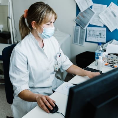 En kvinna i munskydd och läkarrock sitter vid en dator. Hon håller i datorns mus med ena handen.