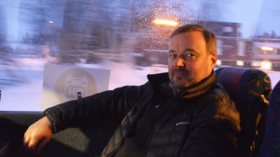 En medelålders man sitter i en buss. Bakom honom synns ett mörkt och snöigt landskap.