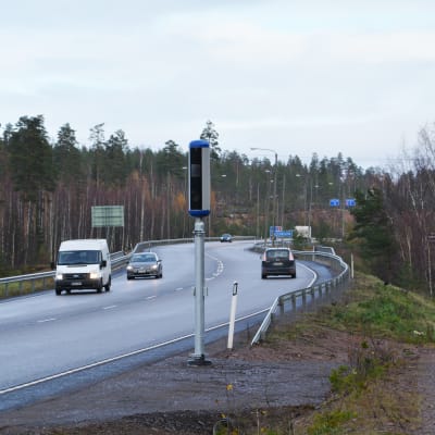 En avlång smal trafikkamera intill en asfalterad väg med några bilar på.