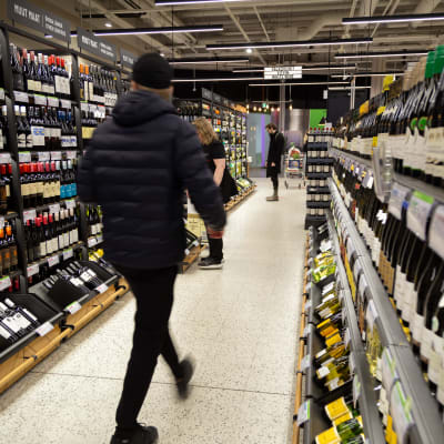 Alkon myymälän sisällä pulloja hyllyissä. Miesasiakas tummansinisessä toppatakissa ja musta pipo päässä on juuri astunut kameran takaa kuvaan reippaasti kävellen hyllyjen välistä käytävää pitkin. Miehen liike on tallentunut epäterävänä.