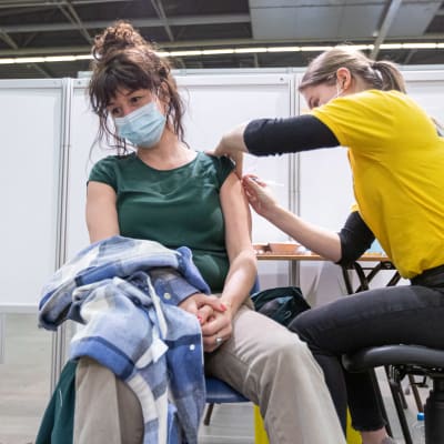 En kvinna i munskydd sitter på en stol. Hon får coronavaccin i ena armen.