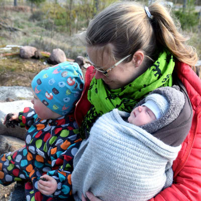 En kvinna sitter utomhus med en sex veckor gamla baby i famnen. Bredvid dem sitter ett två år gammalt barn i en prickig overall.