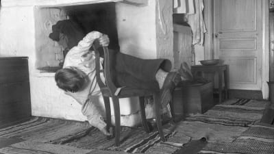 En man leker leken "krypa runt stolen", där han ska vrida sig runt stolen utan att vidröra golvet. Bilden är tagen 1936 i Strömfors, Kungsböle.