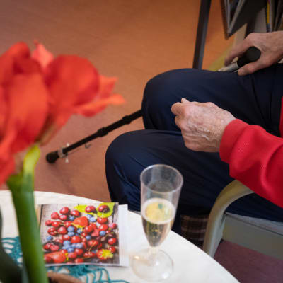 En äldre person sitter med sin promenadkäpp vid ett b ord där det finns ett glas champagne.