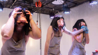 Tre skådespelare i trollaktiga masker och grå förkläden står på en vit scen. 