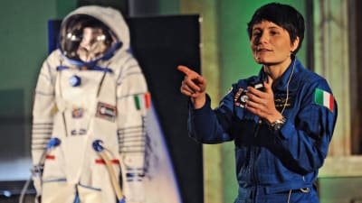 En kvinna klädd i mörkblå overall pekar med ena handen. Till vänster om henne finns en vit rymddräkt.