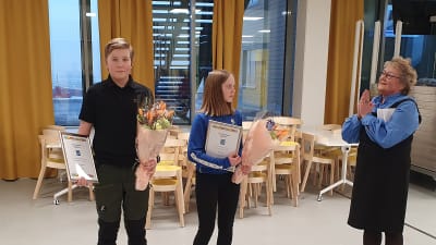 Merja Laaksonen gratulerar Sisu Kymäläinen och Aino Kallio som är duktiga idrottare i Sjundeå. De har diplom och blommor i händerna.